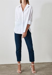 Camisa blanca de botones con mangas enrolladas, combinada con unos vaqueros oscuros ajustados y sandalias blancas de tacón, presentada sobre un fondo neutro.