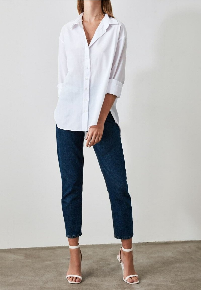 Camisa blanca de botones con mangas enrolladas, combinada con unos vaqueros oscuros ajustados y sandalias blancas de tacón, presentada sobre un fondo neutro.