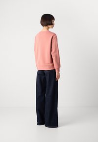 Person with short dark hair stands sideways, wearing a pink sweatshirt and wide-leg dark blue jeans against a plain white background.