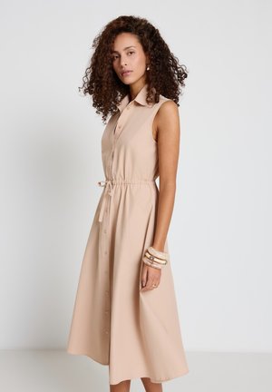 Donna con capelli ricci che indossa un vestito midi beige senza maniche con bottoni, cintura in vita e bracciali spessi al polso.