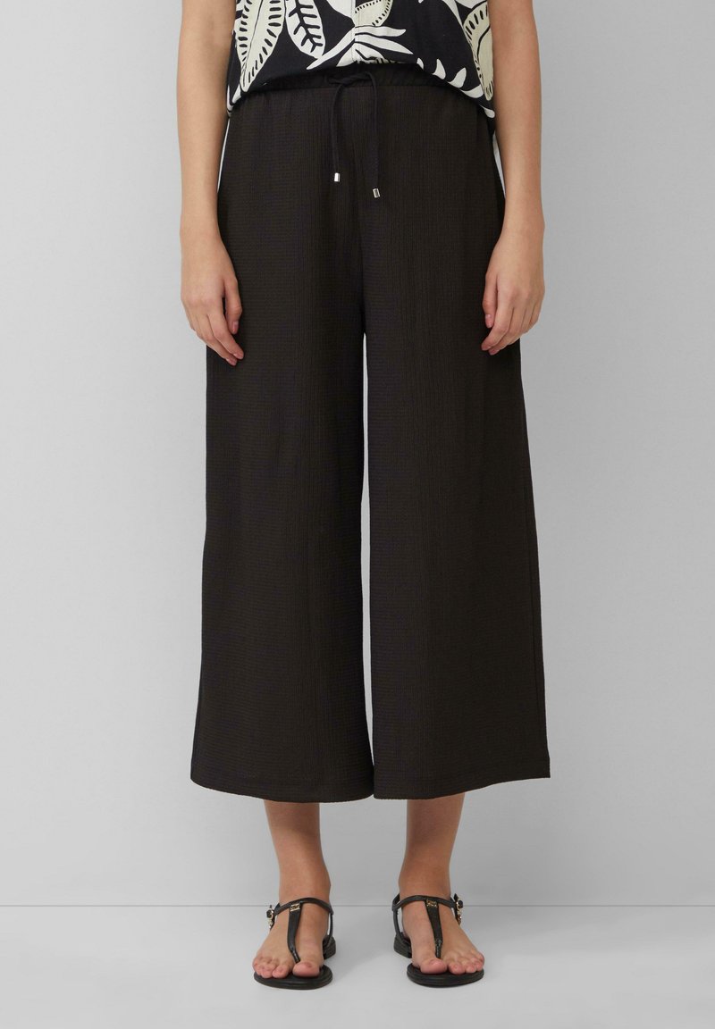 Pantalons larges noirs en tissu texturé, dotés d'une taille élastique et d'un cordon de réglage, associés à des sandales noires simples.
