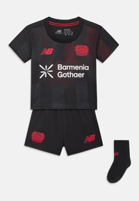 Equipamento de futebol preto com detalhes em vermelho. Camisa de manga curta apresenta um padrão texturizado, o logo do clube e o nome do patrocinador. Inclui calções e uma meia preta.