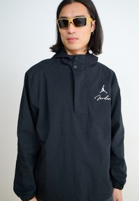 Jachetă neagră cu glugă și guler cu fermoar, având logo-ul alb Jordan. Purtați cu ochelari de soare cu nuanță galbenă, evidențiind un design modern.