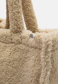 Bolso tote de piel de imitación beige con una superficie texturizada y esponjosa. Presenta asas resistentes y suaves, además de una pequeña etiqueta con logo en el lateral.