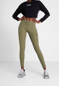 Leggings verde oliva con vita alta, tessuto elasticizzato e logo Puma nero. Abbinati a un crop top nero con branding bianco.