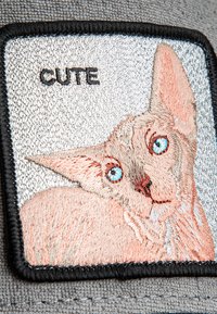 Patch brodé représentant un visage de chat, texture en laine, fourrure pêche, yeux bleus, bord noir, et le mot "MIGNON" en noir au-dessus du design.