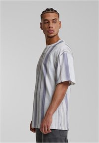 Fonott hajú férfi, laza fehér pólót visel, amelyen függőleges szürke csíkok találhatók, oldalra nézve egy sima háttér előtt.