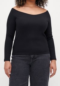 Nainen, jolla on päällään musta olkaimeton pitkähihainen paita ja tummanharmaat farkut, kuvattuna hartioista lantioon yksinkertaista taustaa vasten.