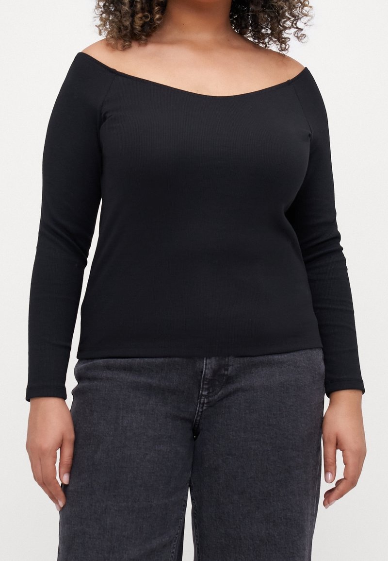 Nainen, jolla on päällään musta olkaimeton pitkähihainen paita ja tummanharmaat farkut, kuvattuna hartioista lantioon yksinkertaista taustaa vasten.