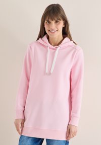 Femme portant un sweat à capuche rose clair avec des cordons blancs et un jean bleu, souriante devant un fond beige uni.