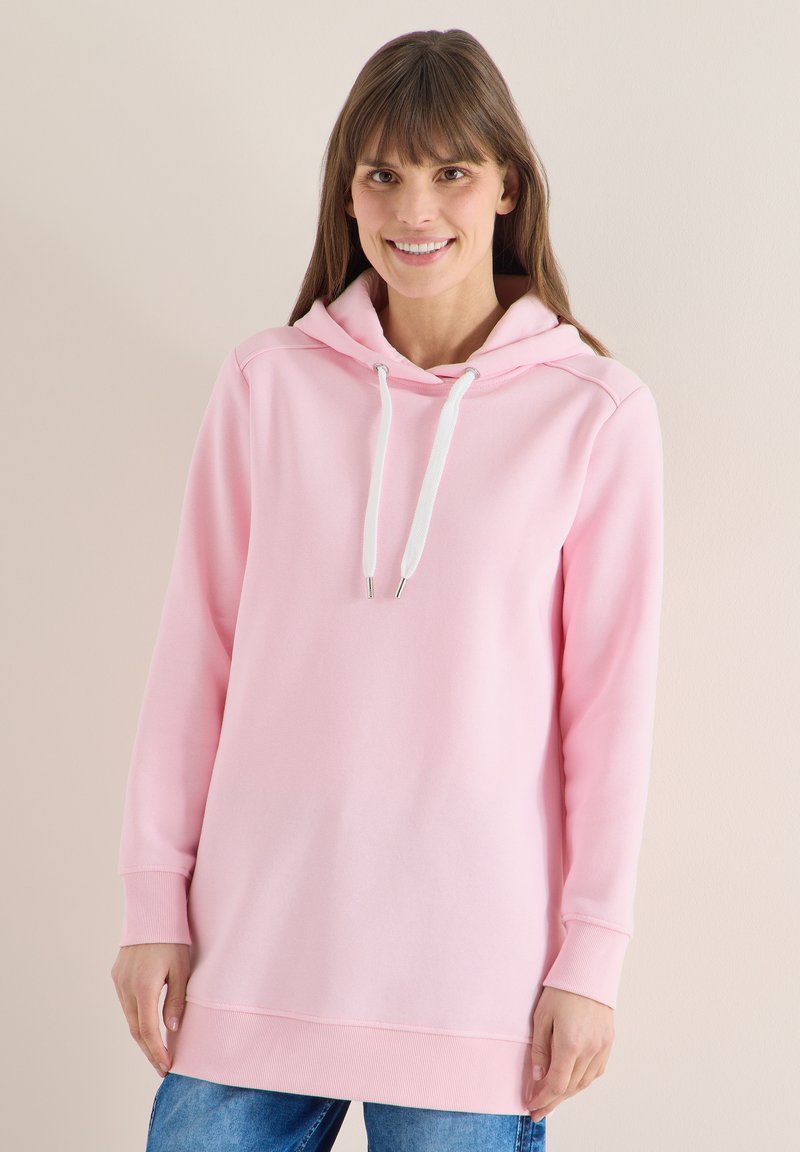 Femme portant un sweat à capuche rose clair avec des cordons blancs et un jean bleu, souriante devant un fond beige uni.