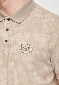 Polo beige con motivo floreale, colletto, bottoni neri e logo EA7 ricamato sul lato sinistro del petto.