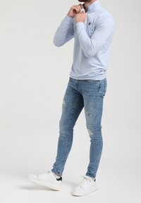 Suéter de manga larga azul claro con un cuello alto y un logo, combinado con unos pantalones vaqueros de denim claro desgastados y zapatillas blancas.