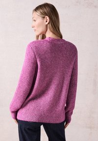 Rab stickad tröja i en livlig rosa färg med ribbat mönster, rund halsringning och långa ärmar, bar tillsammans med mörkblå byxor.