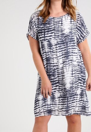 Kobieta ubrana w krótką sukienkę do kolan z czarno-białym wzorem tie-dye, stojąca na jednolitym tle.