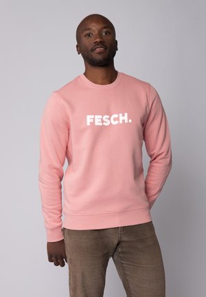 Helles rosa Sweatshirt mit weißem "FESCH." Logo, Rundhalsausschnitt, langen Ärmeln und gerippten Bündchen. Kombiniert mit hellbraunen Hosen.