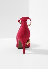 Sandalo alto in suede rosso con cinturino alla caviglia e fibbia, visto da dietro su una superficie e uno sfondo bianchi.