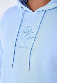 Sudadera de color azul claro hecha de una tela suave, con un bolsillo tipo canguro, cordones con puntas de metal plateado y un logotipo bordado en el pecho.