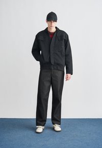 Fekete bomberdzseki cipzárral, két elülső zsebbel, piros csíkos ing fölött viselve. A megjelenést sötét farmernadrág és fehér sportcipő egészíti ki.