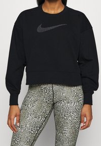 Sweat-shirt noir court avec un swoosh Nike argenté texturé, manches longues et poignets côtelés, associé à un legging vert et noir à motifs.