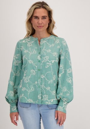 Vrouw in een teal blouse met knopen, versierd met witte bloemenborduursels en volumineuze lange mouwen, gecombineerd met lichtblauwe jeans, staand tegen een witte achtergrond.