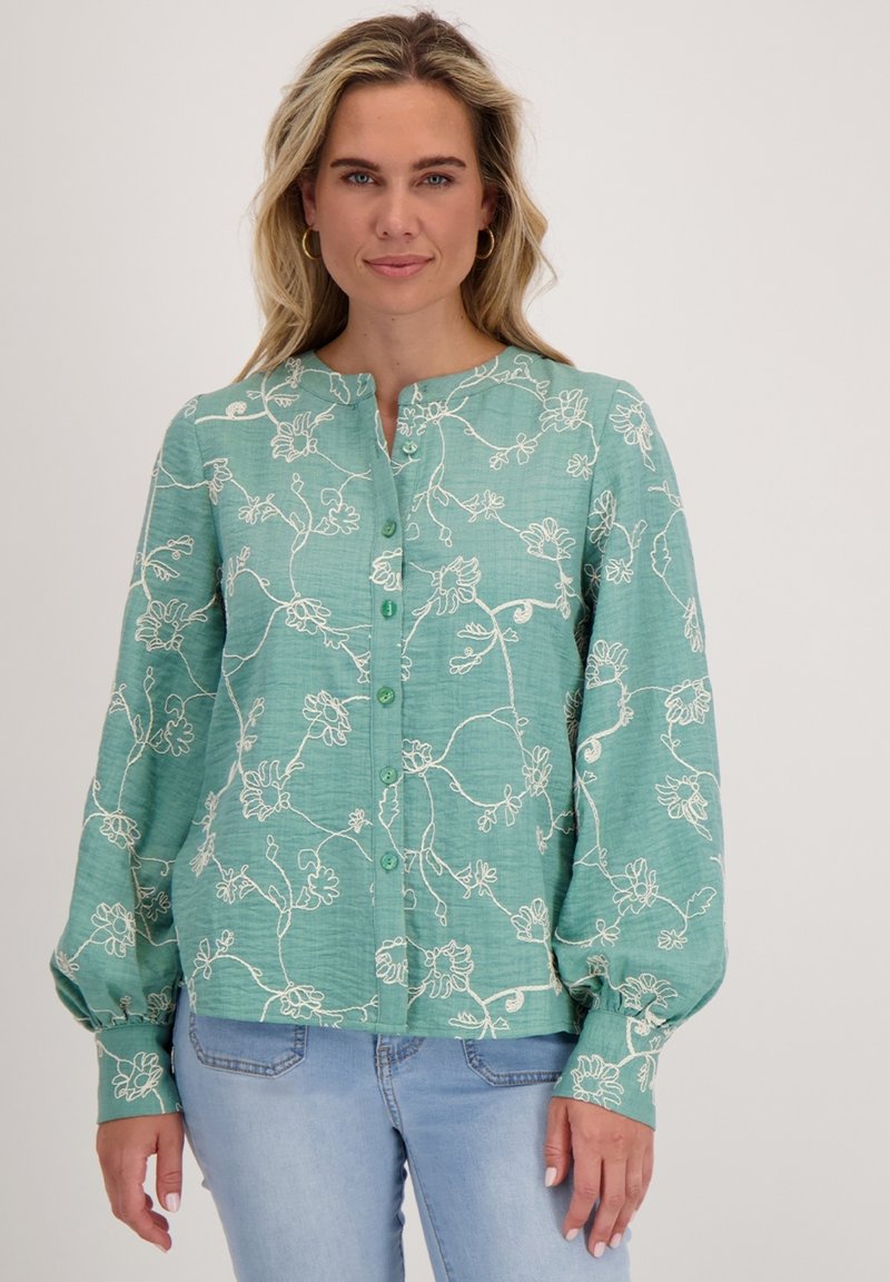 Vrouw in een teal blouse met knopen, versierd met witte bloemenborduursels en volumineuze lange mouwen, gecombineerd met lichtblauwe jeans, staand tegen een witte achtergrond.