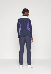 Athletische Jacke mit navyblauer Basis, weißen und lila Akzenten; kombiniert mit navyblauen Hosen und weißen Sneakers. Glattes Material, taillierte Passform.