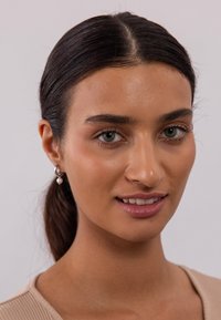 Nahaufnahme einer Person mit mittlerem Hautton und dunklen Haaren, die silberne Creolen mit einem Perlenakzent und ein leichtes, geripptes Top in Beige trägt.