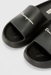 Sorte slidesandaler med åpen tå-design, med en teksturert gummioverdel og logoen "Champion" i hvite bokstaver over stroppen.