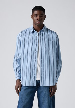 Young man wearing a light blue, vertically striped button-up shirt over a white T-shirt and blue jeans, standing against a plain background.