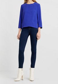 Blusa blu con maniche a tre quarti abbinata a jeans skinny scuri e stivaletti bianchi alla caviglia. Tessuto morbido e design sartoriale.