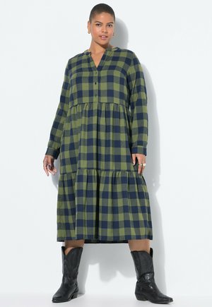 Frau in einem langärmeligen, gestuften Midi-Kleid in grün-blauem Karomuster und schwarzen, bestickten Cowboystiefeln, stehend vor einem weißen Hintergrund.