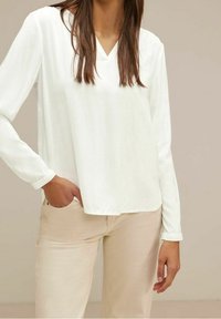 Maglietta bianca a maniche lunghe con scollatura a V, realizzata in tessuto leggero, abbinata a pantaloni beige chiaro. Texture liscia, caduta morbida e vestibilità rilassata.