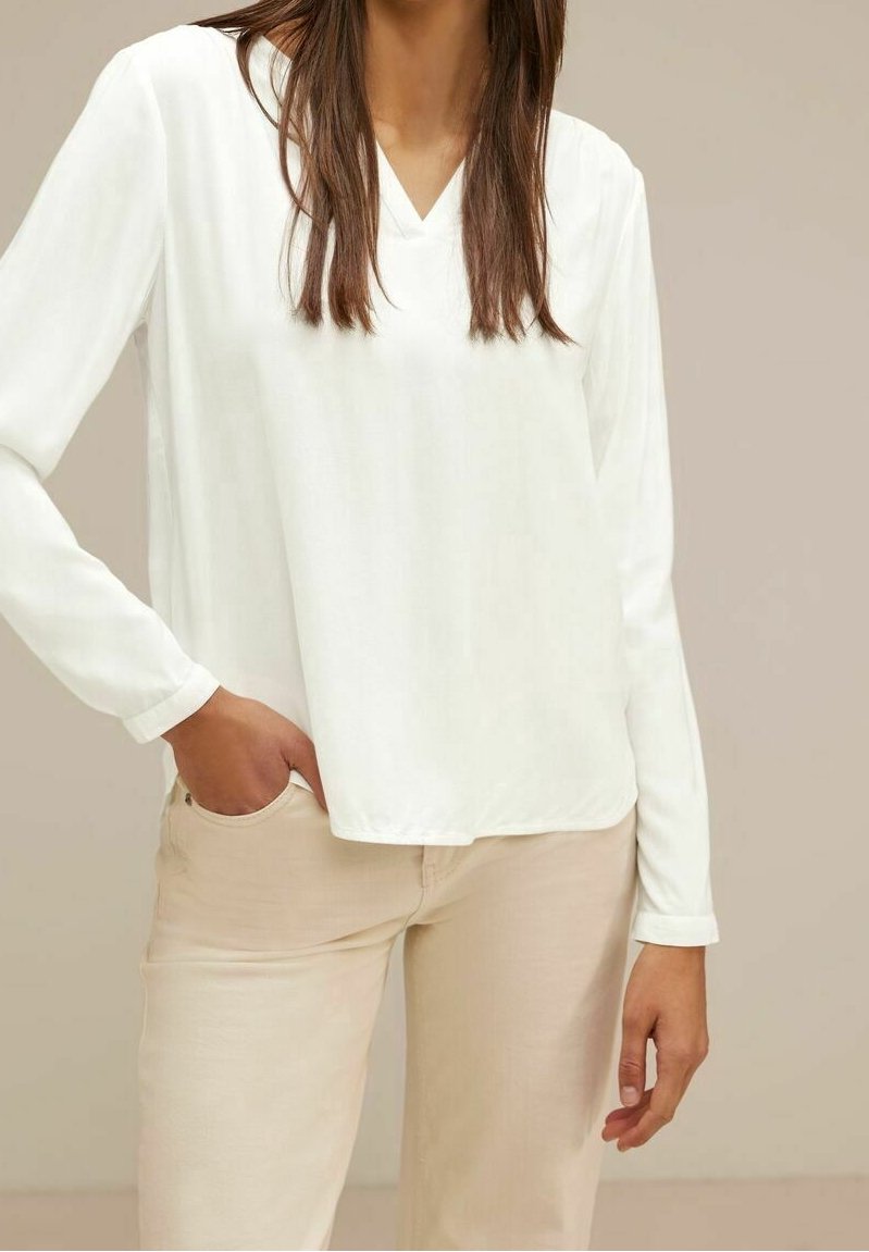 Maglietta bianca a maniche lunghe con scollatura a V, realizzata in tessuto leggero, abbinata a pantaloni beige chiaro. Texture liscia, caduta morbida e vestibilità rilassata.