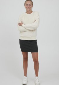 Kremfarget fleecegenser med ribbede mansjetter, kombinert med et svart, tettsittende skjørt. Modellen har på seg hvite joggesko, med armene i kors, foran en ensfarget bakgrunn.