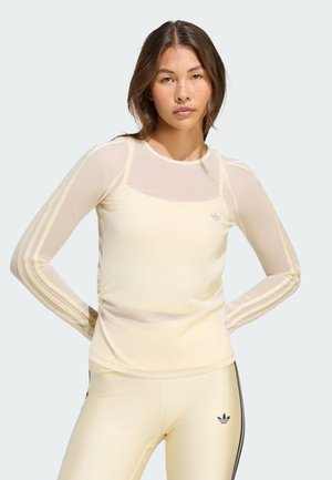 Femme portant un haut transparent à manches longues Adidas jaune pâle et des leggings assortis avec un logo noir sur la hanche, debout devant un fond clair.
