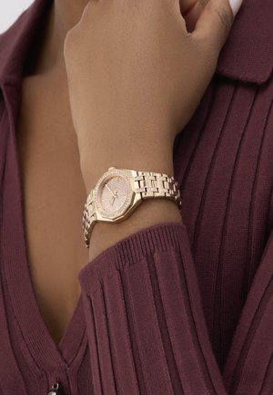 Złoty zegarek z ośmiokątną tarczą, lśniącymi detalami na cyferblacie i metalową bransoletą. Noszony na nadgarstku, obok teksturowanej burgundowej odzieży.