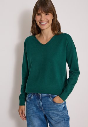Femme souriante aux cheveux bruns portant un pull vert à manches longues et un jean bleu, debout devant un fond clair uni.
