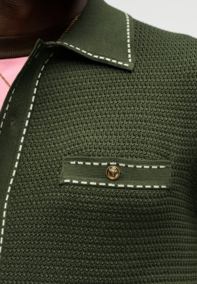 Σκούρο πράσινο πλεκτό πουκάμισο με υφή, με λευκά ραφάκια στο γιακά και στην πλαινή τσέπη, με χρυσό κουμπί με κεφάλι λιονταριού στην πλαινή τσέπη, φορεμένο πάνω από ροζ ένδυμα.