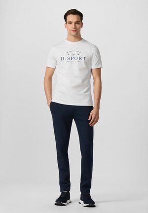 Junger Mann steht frontal, trägt ein weißes H.SPORT T-Shirt, marineblaue Hose und schwarze Sneakers vor einem schlichten grauen Hintergrund.