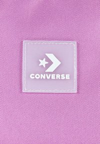 Vijolična teksturirana tkanina z prosojno oznako, na kateri je z belim besedilom "CONVERSE" in dizajnom z zvezdo in puščico.