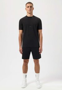 Svart t-shirt med kort ärm och diskret logotyp, tillsammans med svarta shorts. Modellen bär vita strumpor och sneakers, vilket framhäver en enkel atletisk stil.