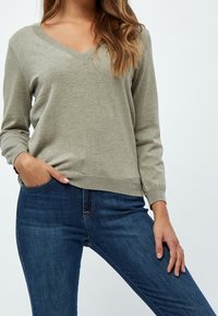 Sweter z dekoltem w serek w kolorze jasnooliwkowym, wykonany z dzianiny, z długimi rękawami i prążkowanym dołem, zestawiony z ciemnoniebieskimi, dopasowanymi dżinsami.