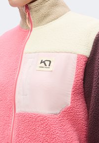 Gros plan d'une veste polaire multicolore avec des sections rose, crème et marron et une poche poitrine rose clair avec un écusson logo Kari Traa.