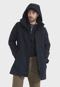 Marineblauer wasserdichter Jacke mit Kapuze, Reißverschluss vorne und Kordelzug. Hat einen entspannten Schnitt und Seitentaschen. Getragen über einem dunklen Pullover.