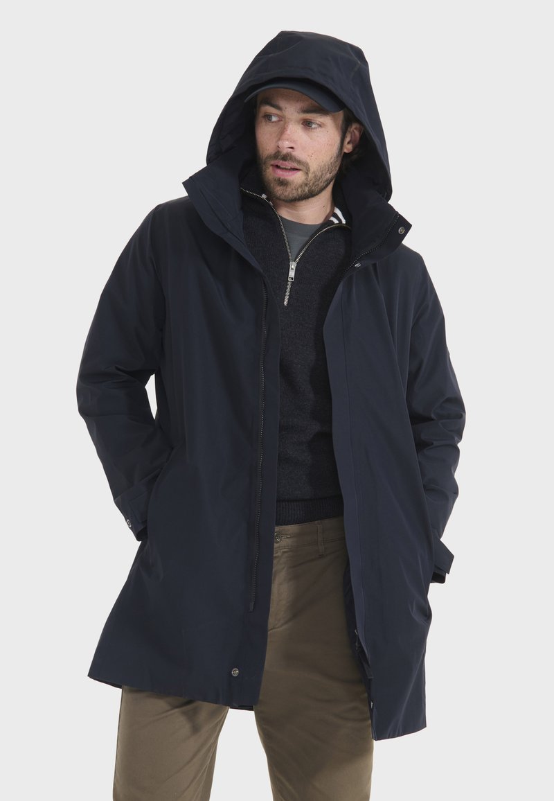 Marineblauer wasserdichter Jacke mit Kapuze, Reißverschluss vorne und Kordelzug. Hat einen entspannten Schnitt und Seitentaschen. Getragen über einem dunklen Pullover.