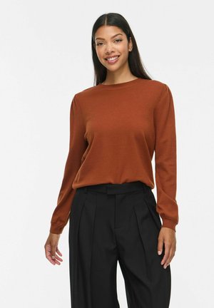 Femme souriante portant un haut à manches longues couleur rouille et un pantalon noir plissé taille haute sur fond uni.