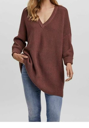 Vrouw draagt een losse, bruingekleurde V-hals trui met textuur, lange mouwen en een blauwe spijkerbroek, met haar hand nonchalant in de trui zak.