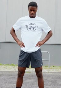 Tricou alb Nike cu logo-ul "Nike Track Club", asociat cu pantaloni scurți gri. Ținuta are o croială lejeră și un material ușor.