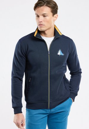 Jeune homme portant une veste zippée bleu marine avec un logo de voilier sur la poitrine et un pantalon bleu, regardant vers le bas à sa gauche.
